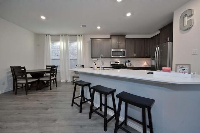 kitchen with stainless steel appliances, a kitchen breakfast bar, dark brown cabinetry, light hardwood / wood-style floors, and decorative backsplash