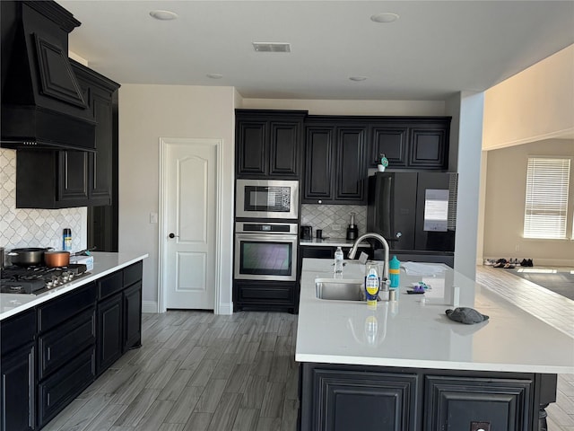 kitchen featuring sink, stainless steel appliances, tasteful backsplash, an island with sink, and custom exhaust hood