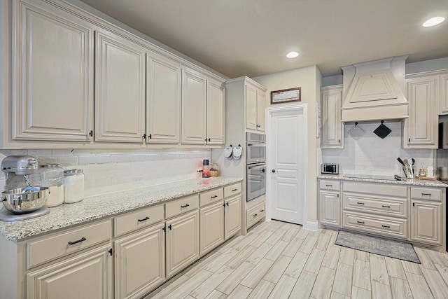 kitchen featuring tasteful backsplash, light stone countertops, custom range hood, black electric cooktop, and stainless steel double oven