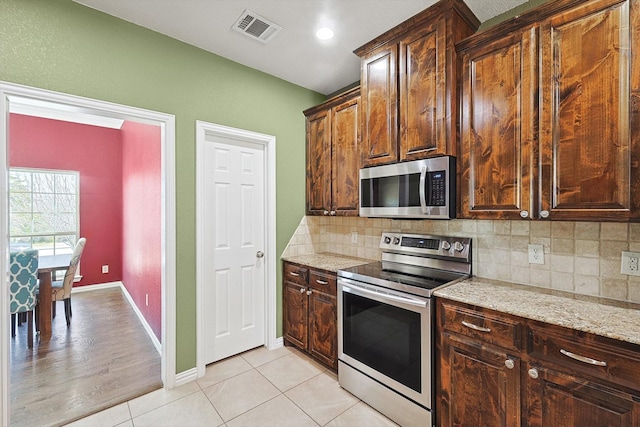 kitchen featuring appliances with stainless steel finishes, dark brown cabinets, light stone counters, light tile patterned flooring, and decorative backsplash