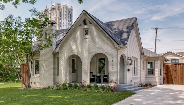 english style home featuring a chimney, a front lawn, roof with shingles, a porch, and brick siding