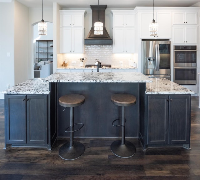 kitchen with stainless steel appliances, premium range hood, white cabinets, and a large island with sink