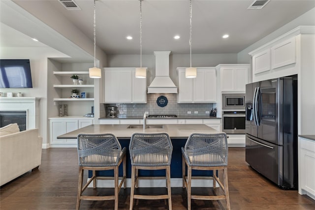 kitchen featuring pendant lighting, appliances with stainless steel finishes, an island with sink, and custom range hood