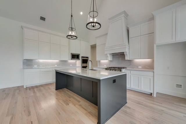 kitchen featuring pendant lighting, stainless steel microwave, custom range hood, an island with sink, and white cabinets