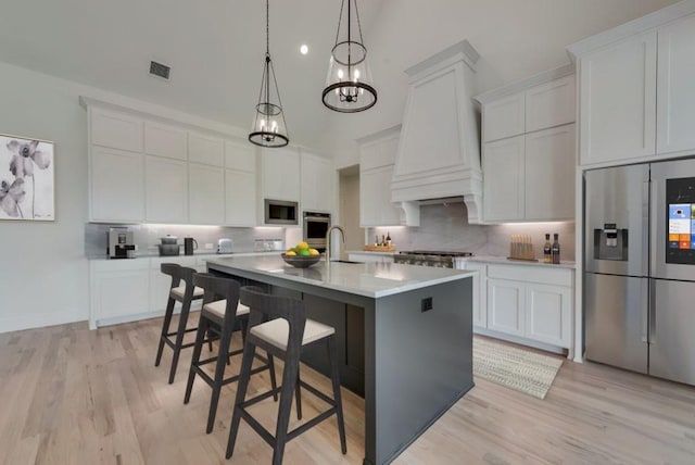 kitchen featuring premium range hood, white cabinetry, decorative light fixtures, appliances with stainless steel finishes, and an island with sink