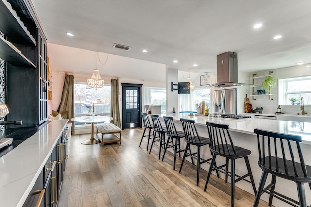 kitchen with stainless steel fridge, tasteful backsplash, island range hood, decorative light fixtures, and light wood-type flooring