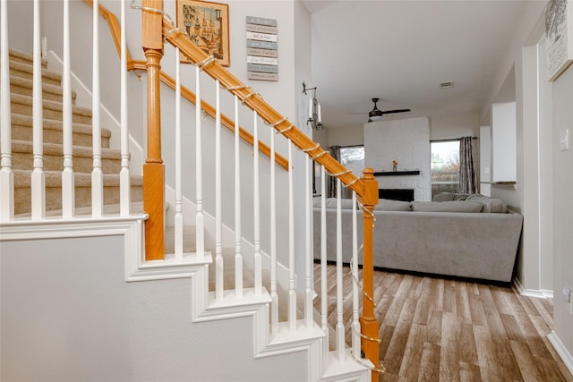 staircase with wood-type flooring, a large fireplace, and ceiling fan