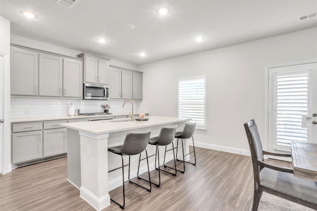 kitchen with a breakfast bar area, gray cabinetry, stainless steel appliances, a center island with sink, and decorative backsplash