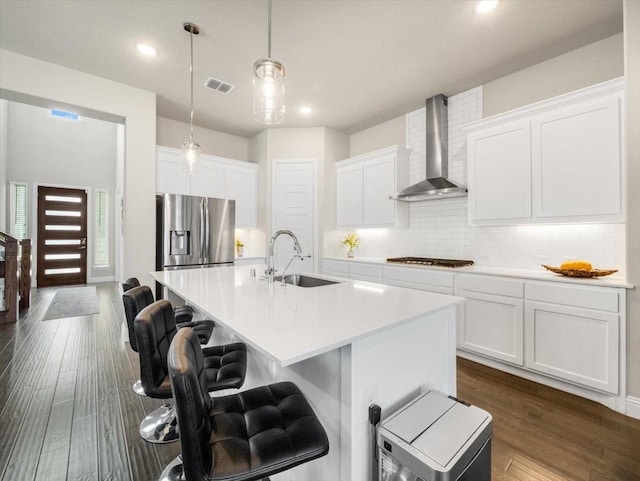 kitchen with hanging light fixtures, stainless steel fridge, an island with sink, and wall chimney exhaust hood