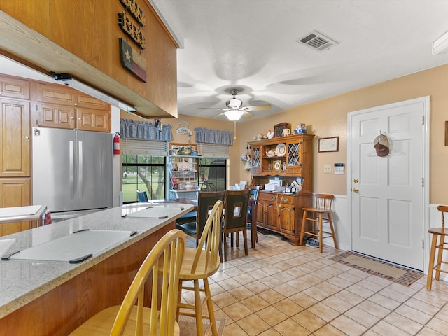 kitchen featuring a kitchen bar, light stone counters, light tile patterned floors, stainless steel refrigerator, and ceiling fan