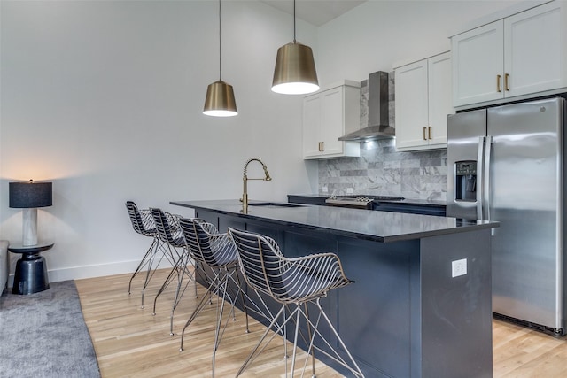 kitchen featuring dark countertops, wall chimney exhaust hood, decorative light fixtures, white cabinetry, and stainless steel refrigerator with ice dispenser