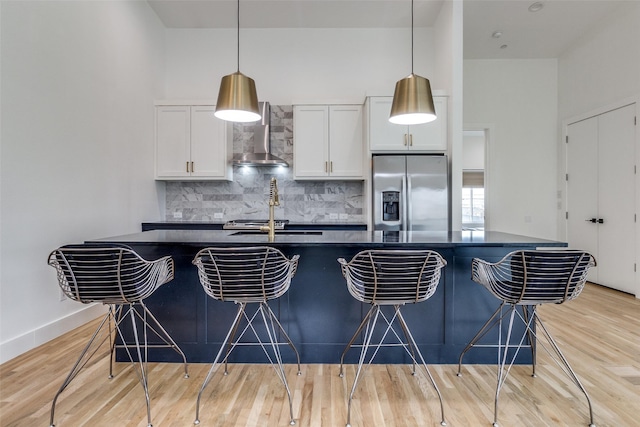 kitchen featuring dark countertops, wall chimney range hood, stainless steel refrigerator with ice dispenser, and white cabinets