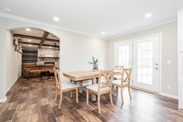 dining area featuring crown molding, dark wood-type flooring, ceiling fan, a large fireplace, and beamed ceiling