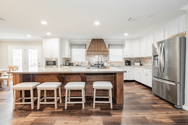 kitchen featuring stainless steel appliances, custom range hood, white cabinets, and a center island with sink