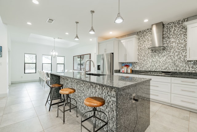 kitchen with wall chimney exhaust hood, white cabinetry, dark stone countertops, a large island with sink, and stainless steel appliances