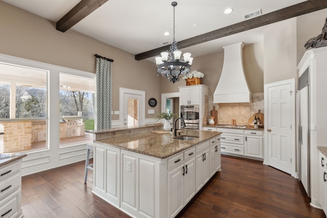 kitchen featuring sink, premium range hood, white cabinetry, a center island with sink, and decorative backsplash