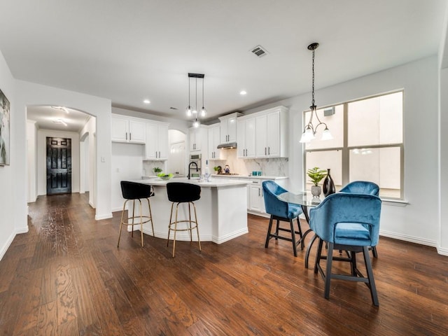 kitchen with dark wood-type flooring, a breakfast bar, a center island with sink, hanging light fixtures, and white cabinets