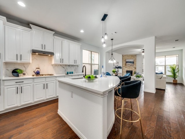 kitchen featuring dark wood-type flooring, white cabinetry, tasteful backsplash, a center island with sink, and a stone fireplace