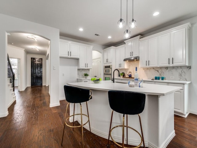 kitchen featuring dark wood-type flooring, hanging light fixtures, a center island with sink, decorative backsplash, and white cabinets