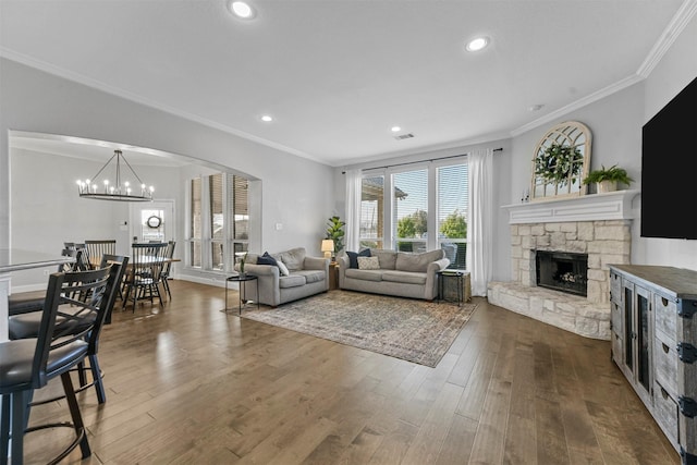 living area featuring dark wood-type flooring, crown molding, a stone fireplace, and an inviting chandelier