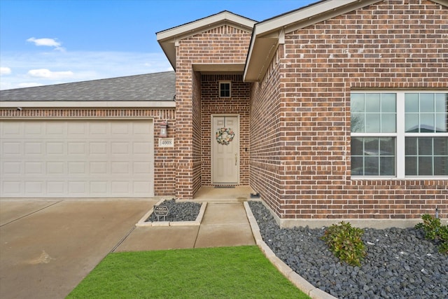 doorway to property with an attached garage, roof with shingles, concrete driveway, and brick siding