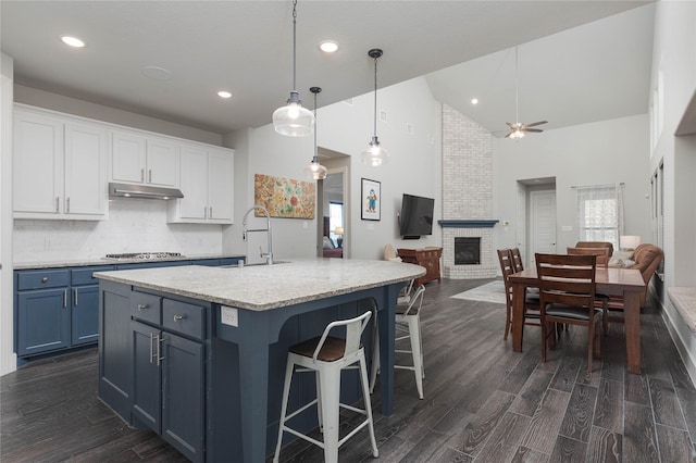 kitchen with dark wood-type flooring, white cabinetry, hanging light fixtures, a kitchen island with sink, and a brick fireplace