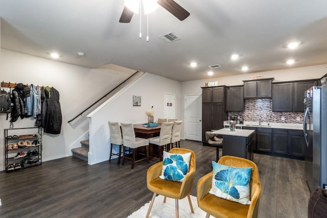 kitchen featuring dark wood-type flooring, dark brown cabinetry, stainless steel refrigerator, a kitchen island with sink, and decorative backsplash