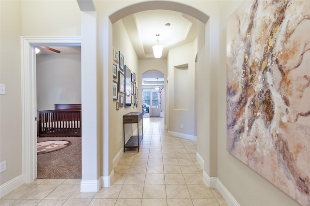 hallway featuring light carpet, light tile patterned floors, baseboards, arched walkways, and a tray ceiling