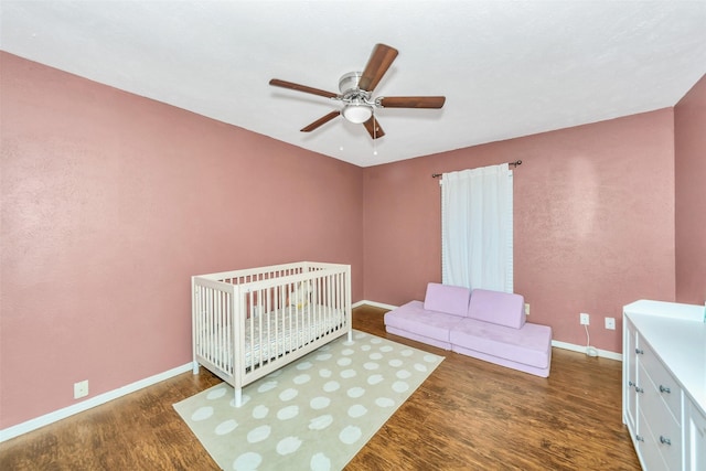 bedroom with a nursery area, dark wood-type flooring, and ceiling fan