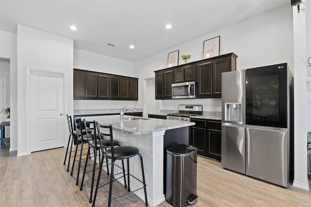 kitchen with sink, a kitchen island with sink, stainless steel appliances, dark brown cabinets, and light hardwood / wood-style flooring