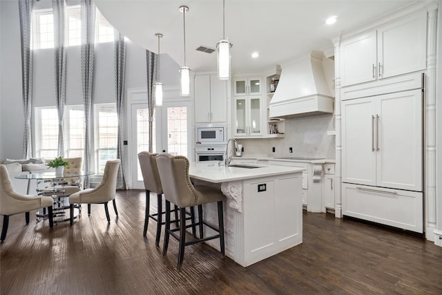 kitchen featuring sink, custom exhaust hood, white cabinetry, a center island with sink, and white microwave