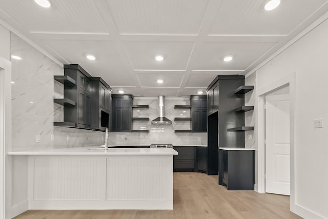 kitchen with wall chimney range hood, light hardwood / wood-style flooring, tasteful backsplash, coffered ceiling, and kitchen peninsula