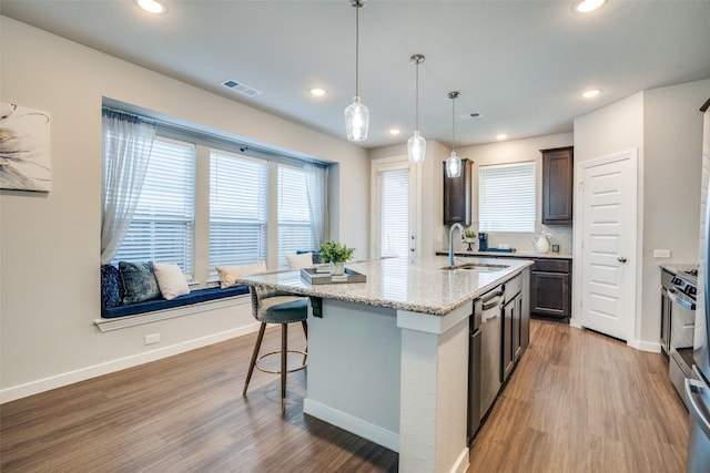 kitchen featuring sink, decorative light fixtures, dark brown cabinets, a center island with sink, and light stone countertops