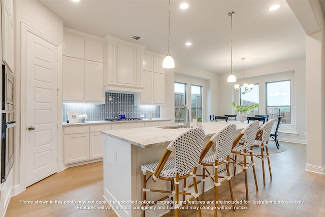 kitchen featuring pendant lighting, an island with sink, sink, white cabinets, and light stone countertops