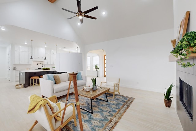 living room with high vaulted ceiling, a fireplace, sink, ceiling fan, and light hardwood / wood-style flooring