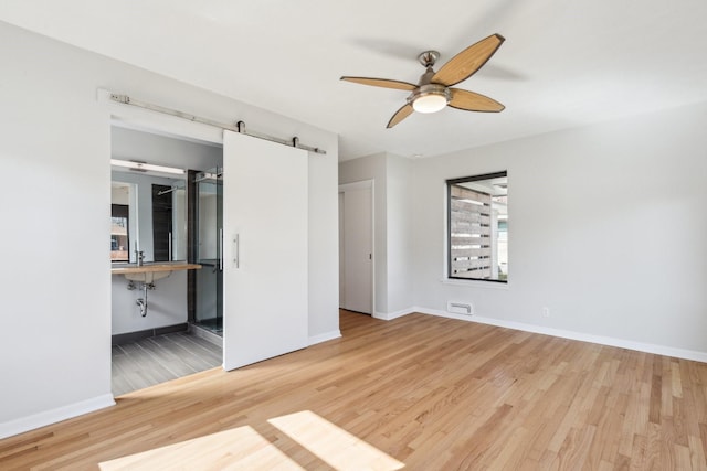 unfurnished bedroom with ceiling fan, ensuite bathroom, a barn door, and light wood-type flooring