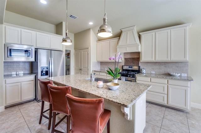 kitchen with appliances with stainless steel finishes, white cabinets, custom exhaust hood, hanging light fixtures, and a kitchen island with sink