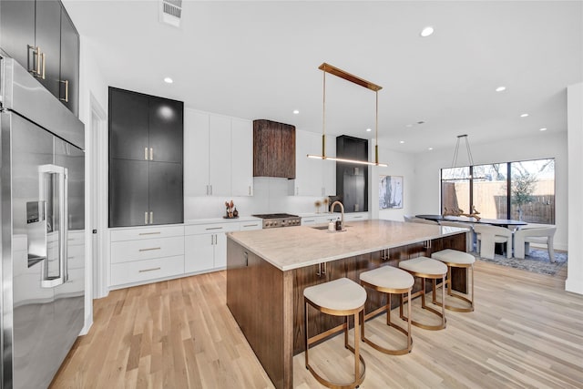 kitchen with stainless steel built in refrigerator, an island with sink, hanging light fixtures, and light wood-type flooring