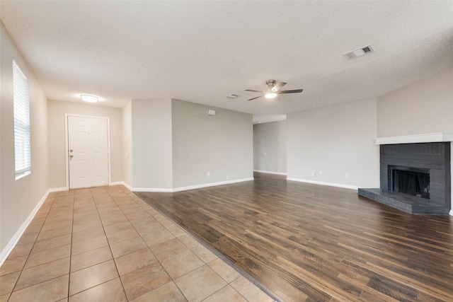 unfurnished living room with ceiling fan, a textured ceiling, wood finished floors, visible vents, and a brick fireplace