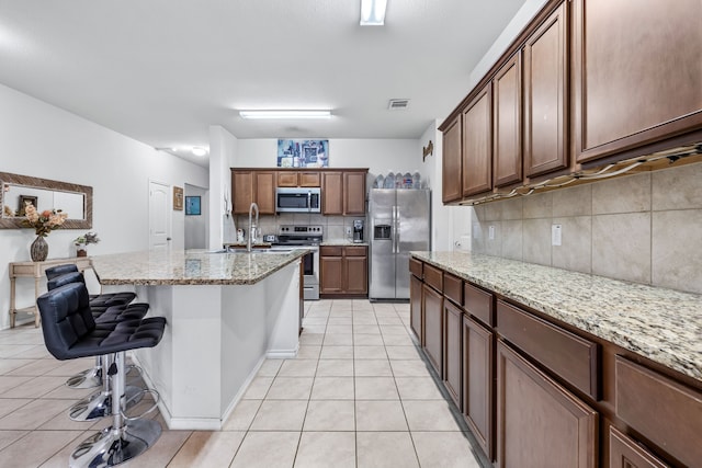 kitchen featuring light tile patterned flooring, a center island with sink, appliances with stainless steel finishes, a kitchen breakfast bar, and light stone countertops