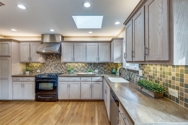 kitchen featuring wall chimney exhaust hood, sink, a skylight, black gas range oven, and light hardwood / wood-style floors