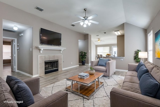 living room featuring sink, ceiling fan, a tiled fireplace, vaulted ceiling, and light wood-type flooring