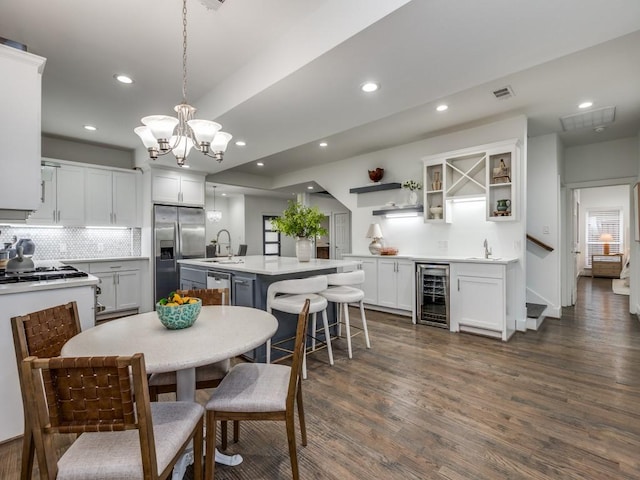 kitchen featuring decorative light fixtures, an island with sink, white cabinets, beverage cooler, and stainless steel appliances