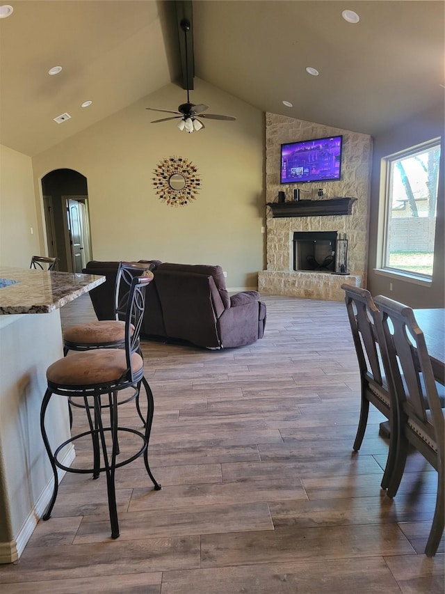 living room featuring hardwood / wood-style flooring, ceiling fan, a stone fireplace, and lofted ceiling with beams