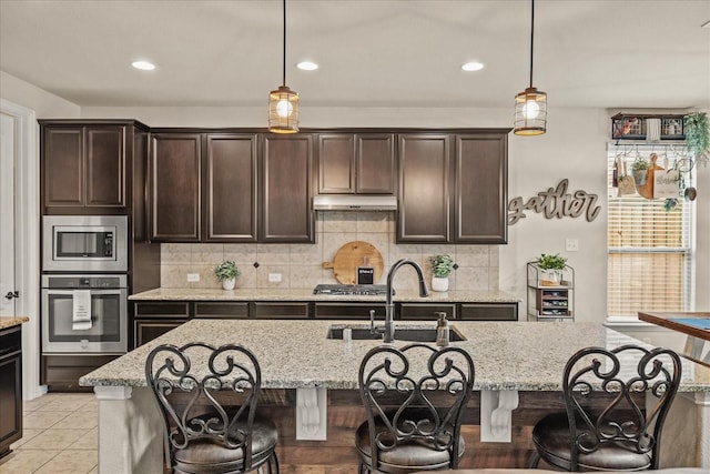 kitchen with sink, a kitchen island with sink, stainless steel appliances, light stone counters, and dark brown cabinetry