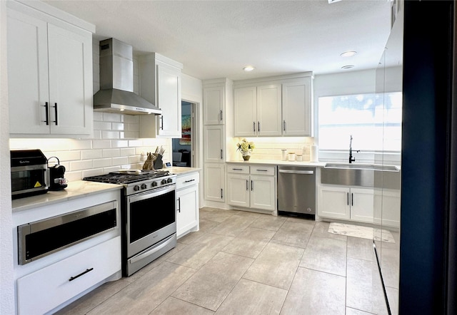 kitchen with stainless steel appliances, light countertops, white cabinets, a sink, and wall chimney exhaust hood