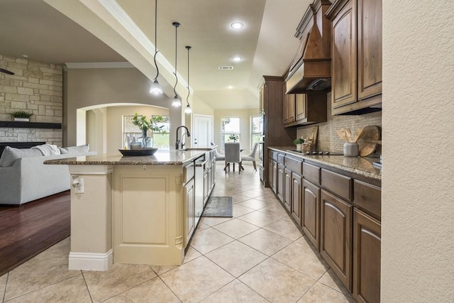 kitchen featuring custom exhaust hood, hanging light fixtures, an island with sink, light stone countertops, and decorative backsplash