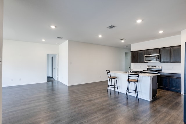 kitchen with dark wood-type flooring, dark brown cabinets, stainless steel appliances, a kitchen breakfast bar, and a center island with sink