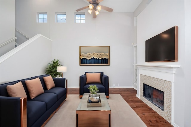 living room with ceiling fan, wood-type flooring, a tiled fireplace, and a high ceiling