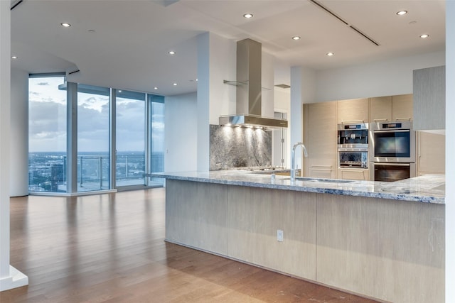 kitchen with ventilation hood, tasteful backsplash, light stone counters, stainless steel double oven, and light wood-type flooring